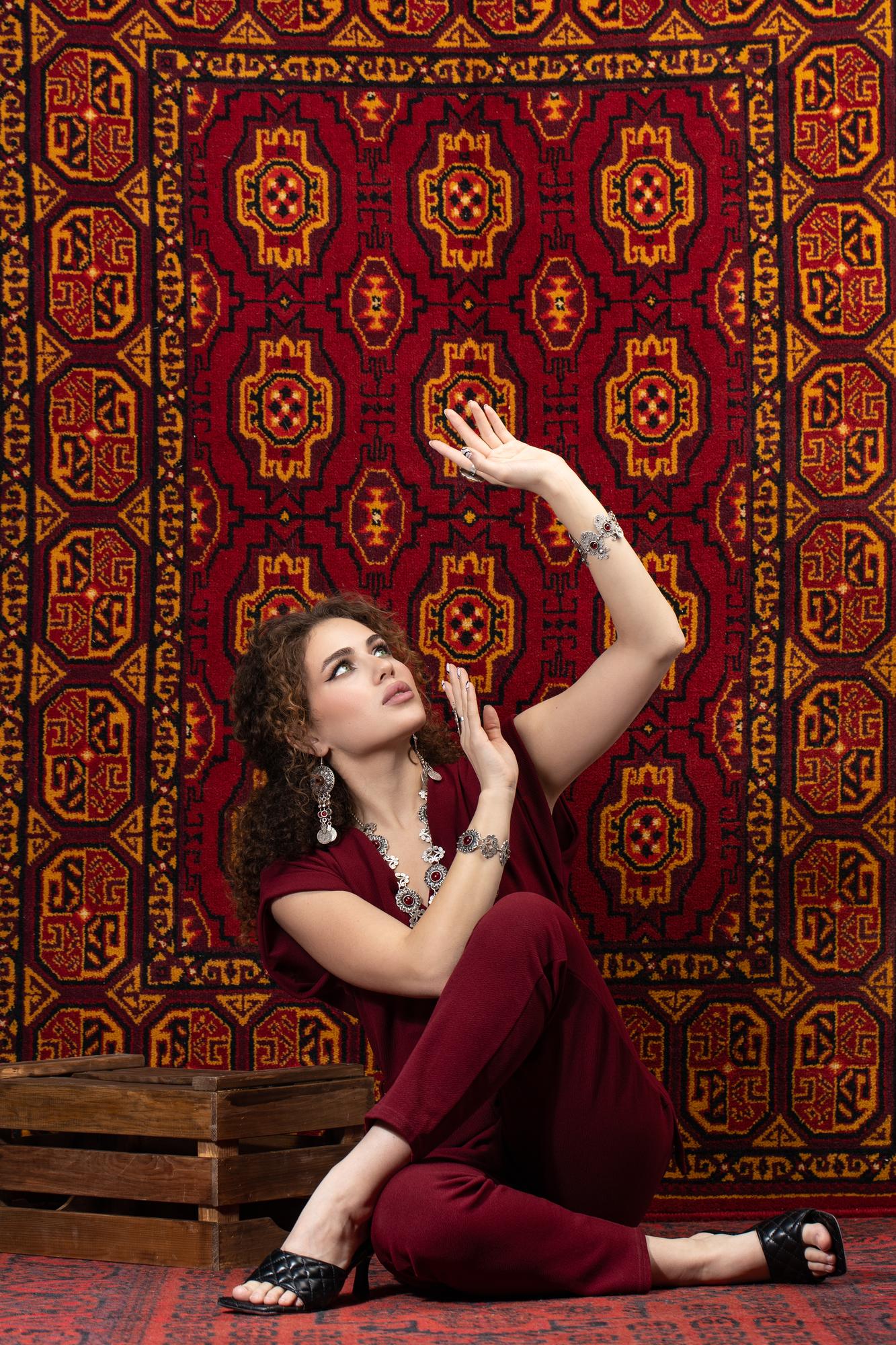 Curly-haired woman in maroon, adorned with intricate silver jewelry, poses elegantly on a rich red and gold traditional rug backdrop.